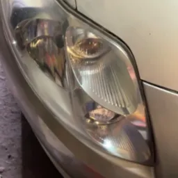 Reflektor samochodu w kolorze srebrnym, widoczny z bliska, z wyraźnymi detalami klosza. Część karoserii w tle. Zdjęcie z warsztatu.