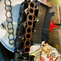 Robert Robert - Demontaż kolektora ssącego z uszczelkami i czujnikiem temperatury na warsztatowym stole. Widoczne ślady użytkowania i elementy otoczenia warsztatowego.