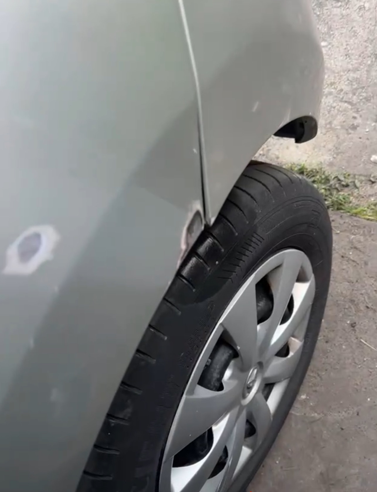 Fragment srebrnego auta z widocznymi uszkodzeniami karoserii i śladami szpachlowania przy kole. Widoczny fragment opony i felgi.