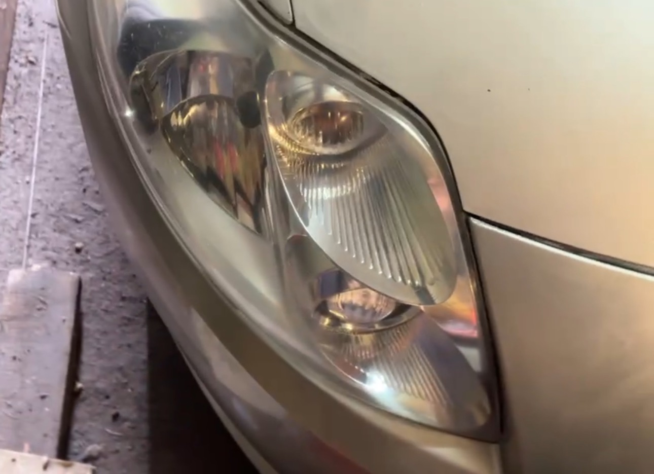 Reflektor samochodu w kolorze srebrnym, widoczny z bliska, z wyraźnymi detalami klosza. Część karoserii w tle. Zdjęcie z warsztatu.