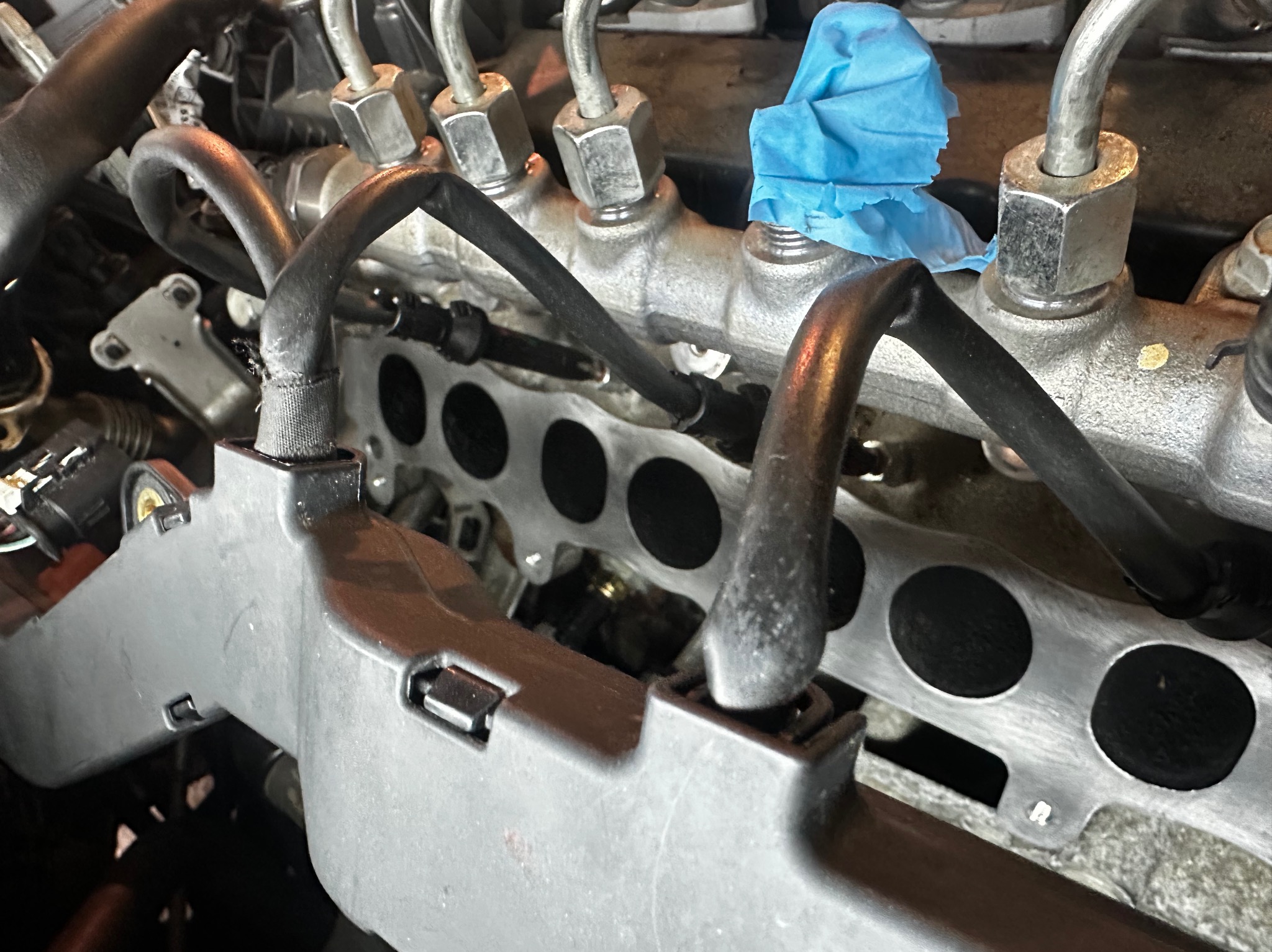 Silnik samochodowy w trakcie naprawy: widoczne wtryskiwacze, przewody paliwowe i kolektor ssący. Fragment osłonięty niebieską taśmą. Detal mechaniki pojazdowej.