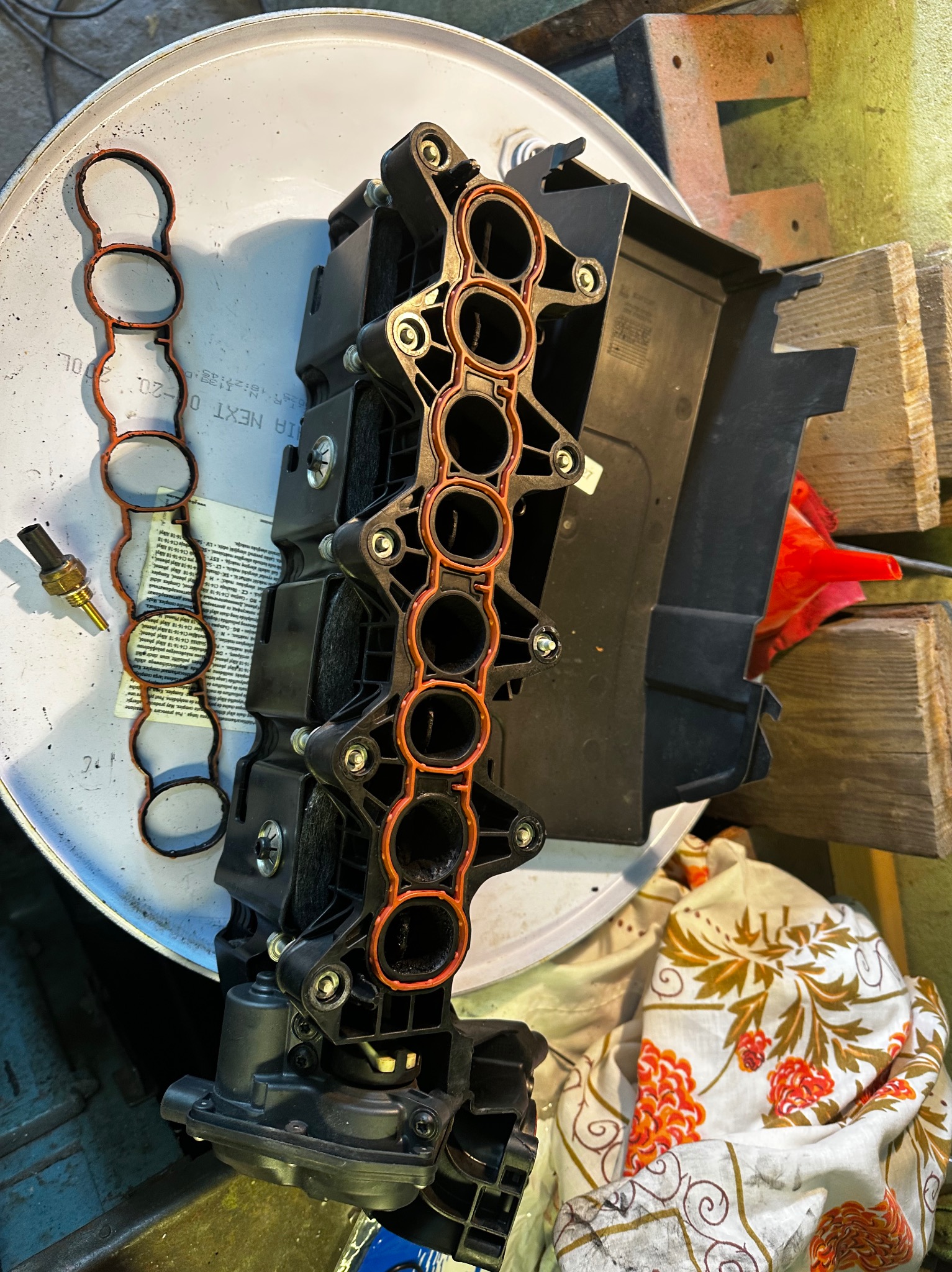 Demontaż kolektora ssącego z uszczelkami i czujnikiem temperatury na warsztatowym stole. Widoczne ślady użytkowania i elementy otoczenia warsztatowego.