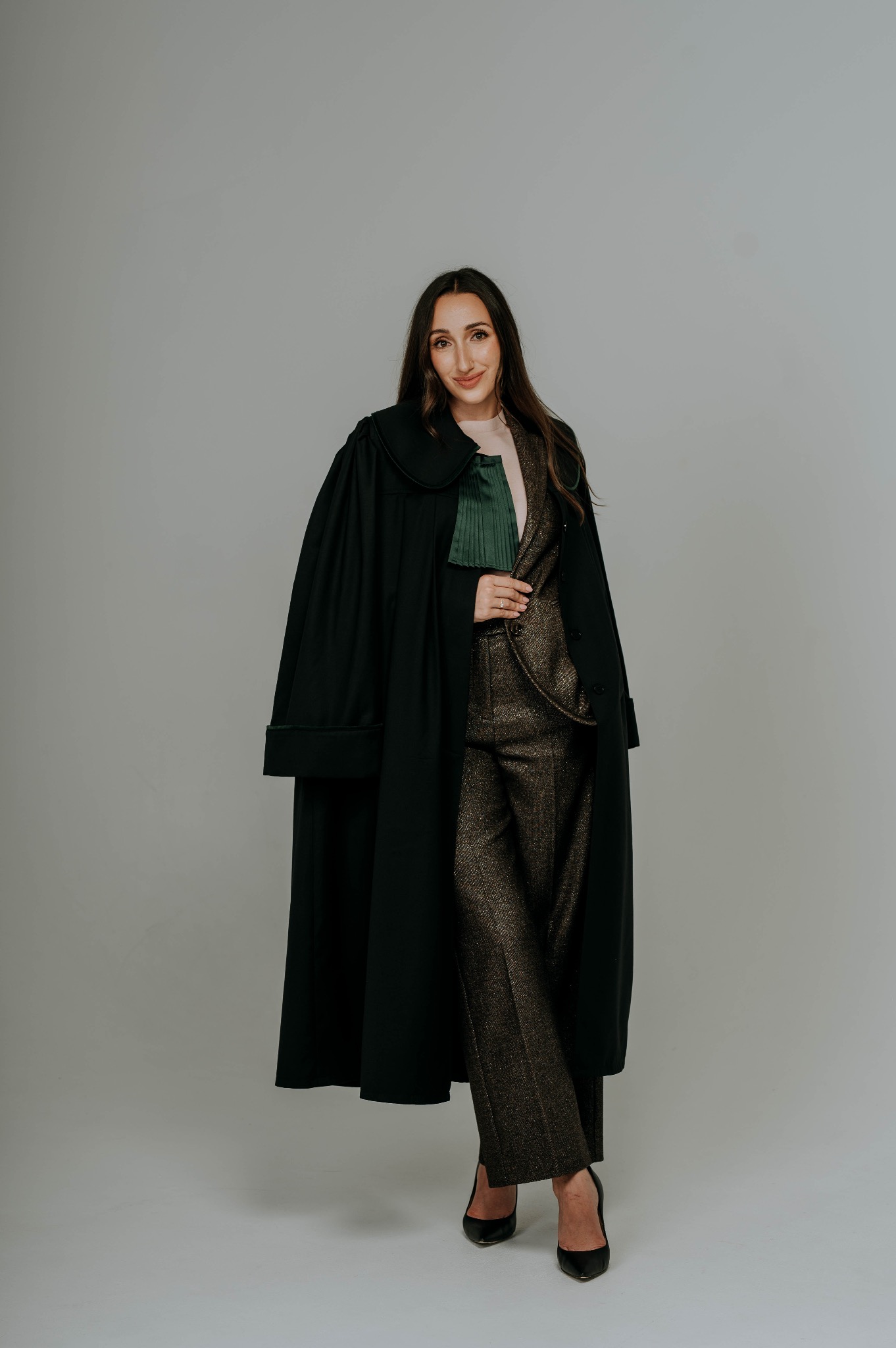 Uśmiechnięta kobieta w todze adwokackiej i eleganckim garniturze, na neutralnym tle. Widoczny zielony żabot i czarne szpilki. Całość w stonowanej kolorystyce.