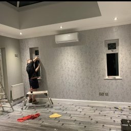 Remontowy Armagedon - Pracownik montuje tapetę w pomieszczeniu z oknami i klimatyzacją. Na podłodze folia ochronna, widoczne narzędzia pracy i drabina.