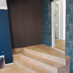 Remontowy Armagedon - Nowoczesne wnętrze z drewnianymi schodami, panelami ściennymi i tapetą w stylu art deco. Widok na białe drzwi i dalsze pomieszczenie z drewnianą podłogą.