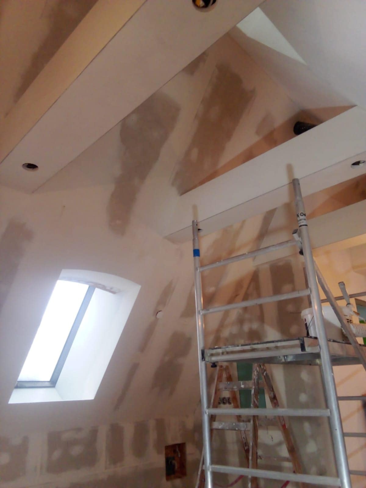 Poddasze w trakcie prac wykończeniowych: szpachlowane ściany, okno dachowe, aluminiowa drabina. Widoczne niedoskonałości przed malowaniem.