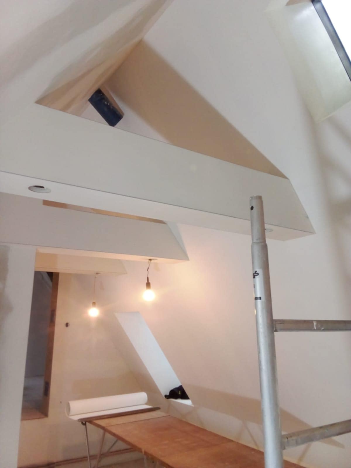 Skośne poddasze w trakcie prac wykończeniowych, widoczne rusztowanie, lampy wiszące i przygotowane materiały. Nietypowy kształt sufitu.