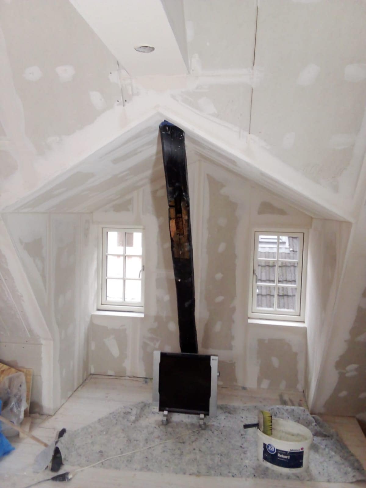 Poddasze ze skośnymi ścianami w trakcie prac. Widoczne okna, fragment instalacji i ślady szpachlowania na ścianach. Na podłodze materiał izolacyjny i wiadro z narzędziami.