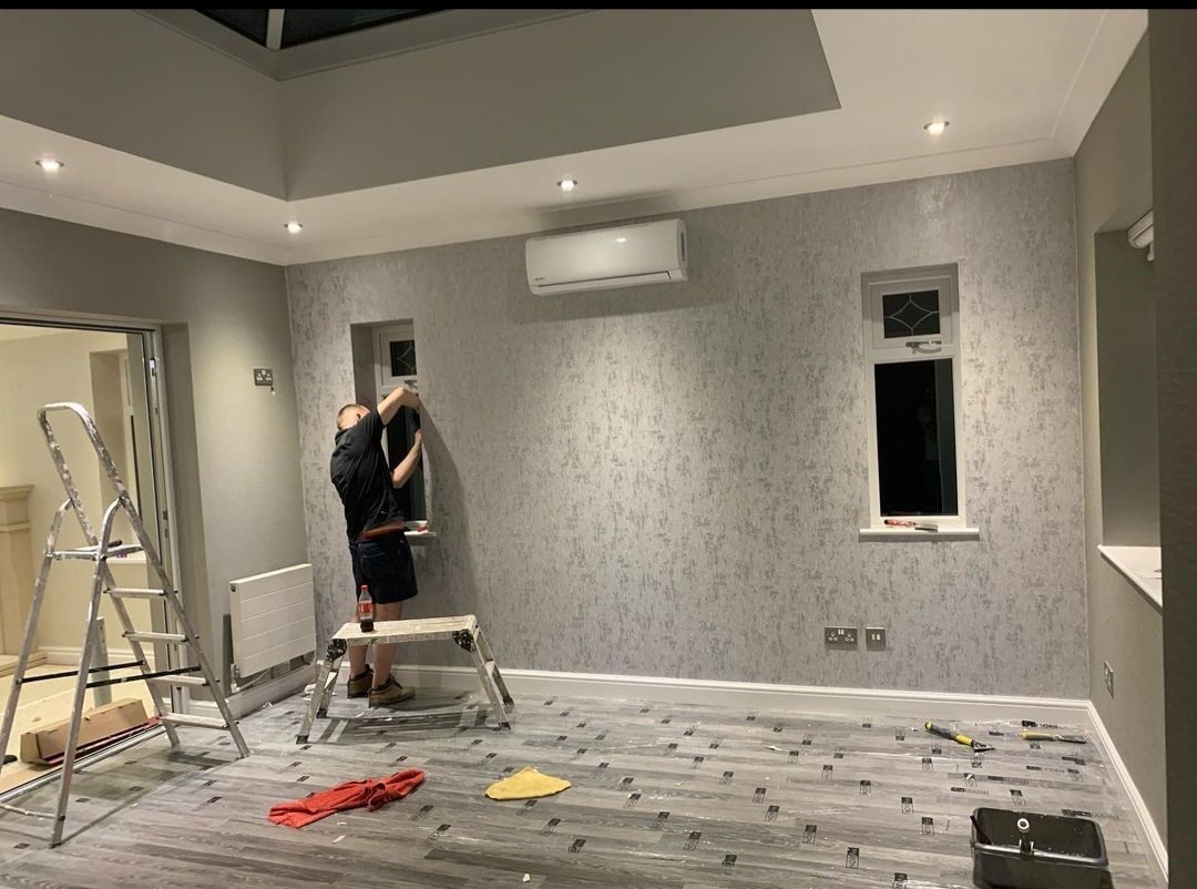 Pracownik montuje tapetę w pomieszczeniu z oknami i klimatyzacją. Na podłodze folia ochronna, widoczne narzędzia pracy i drabina.