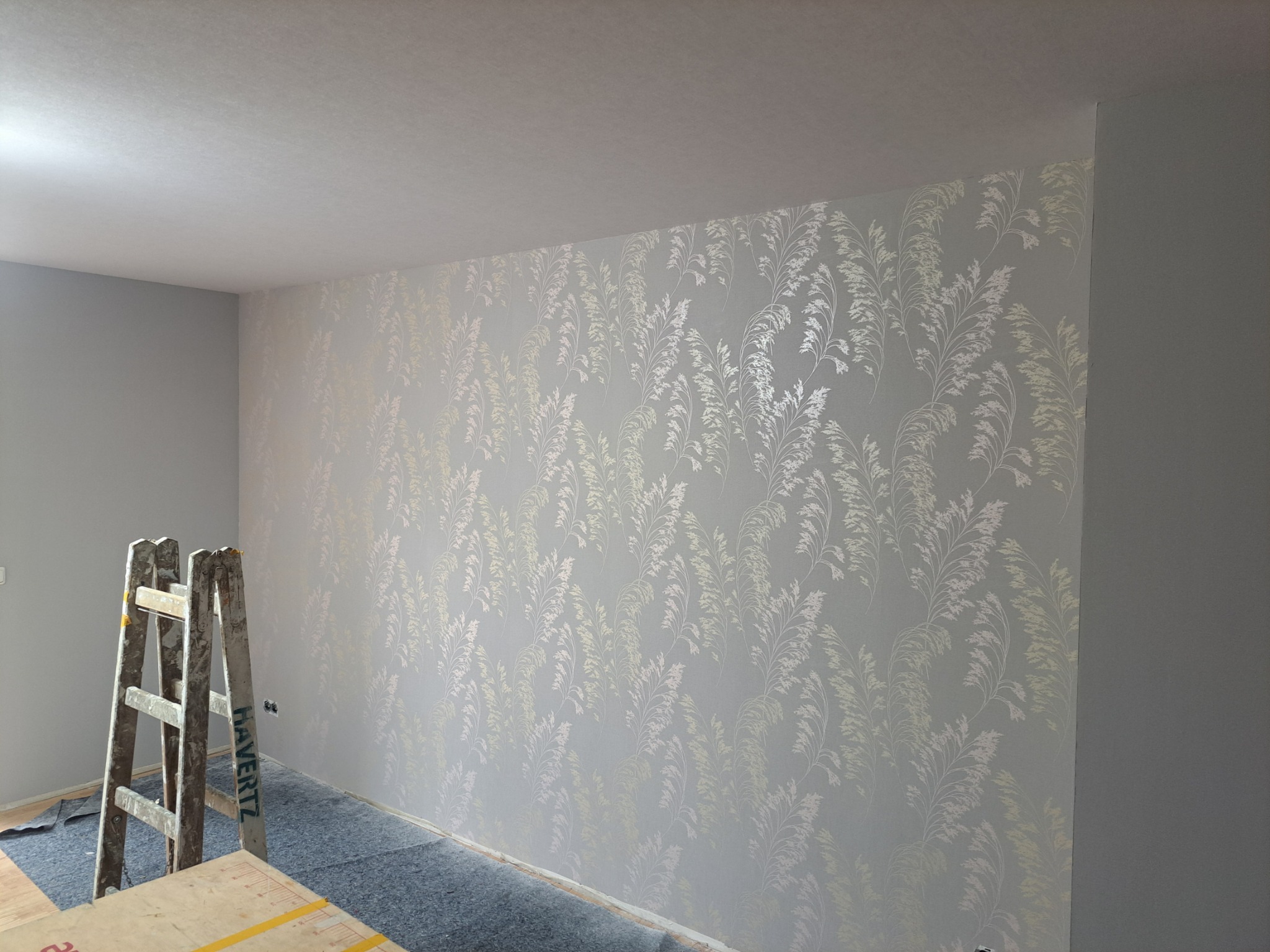 Pokój po tapetowaniu z wzorem roślinnym, widoczna drabina malarska i zabezpieczona podłoga. Ściana pomalowana na szaro. Wykończenie wnętrza.