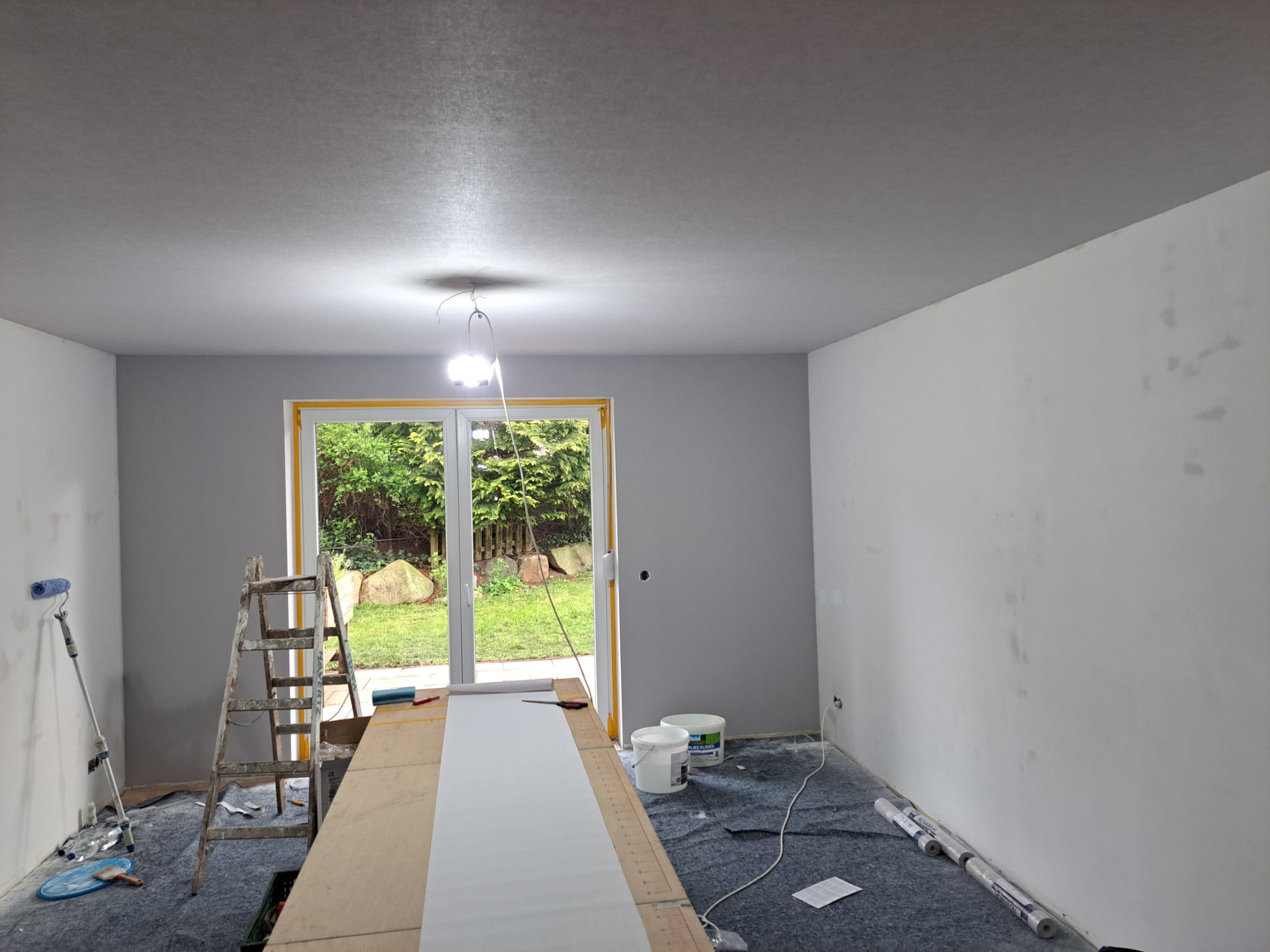 Wnętrze pokoju w trakcie malowania: ściany częściowo szare, częściowo białe, drabina, wałek malarski, zabezpieczona podłoga. Widok na ogród przez przeszklone drzwi.