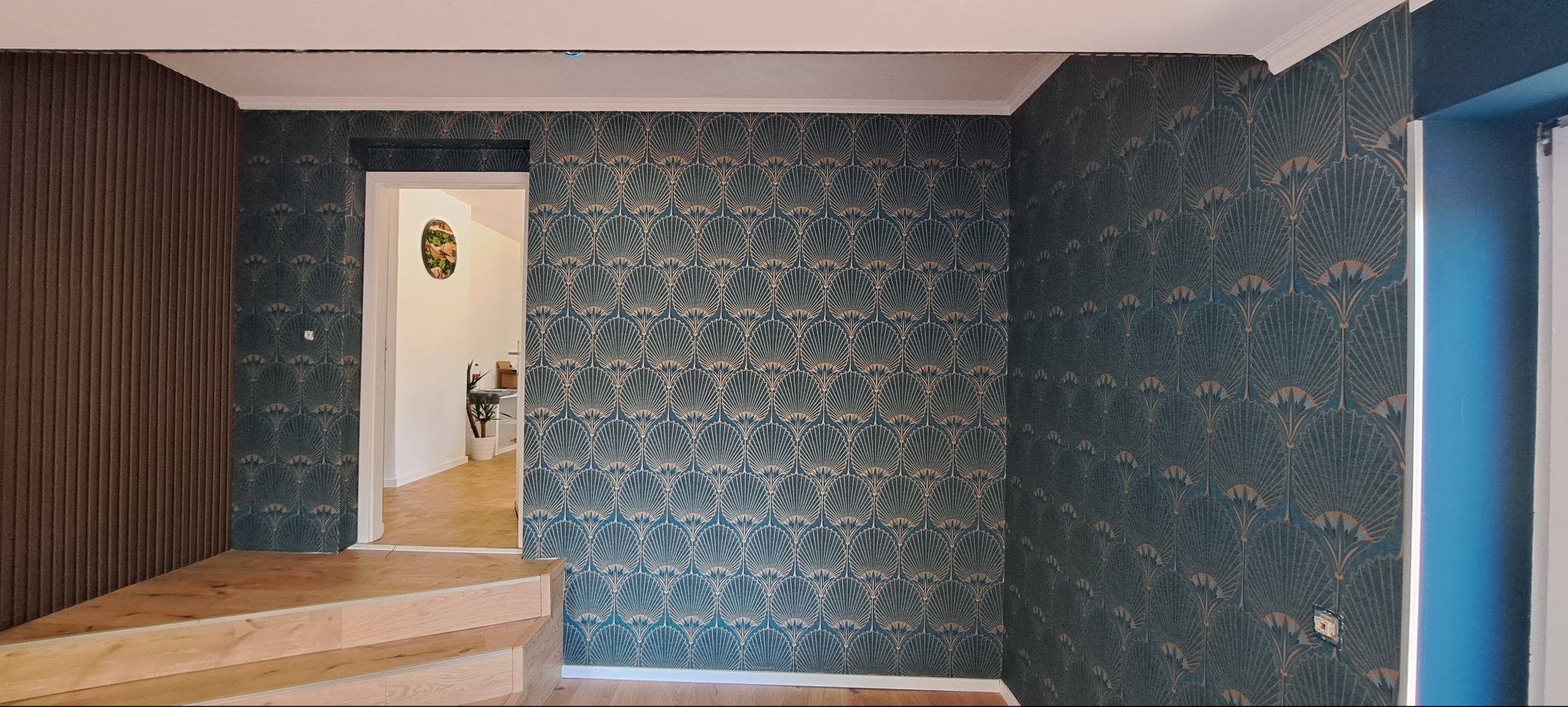 Wykończone wnętrze: ściana w dekoracyjnej, turkusowo-złotej tapecie w geometryczny wzór wachlarzy, drewniane panele ścienne i widok na jasne pomieszczenie przez drzwi.