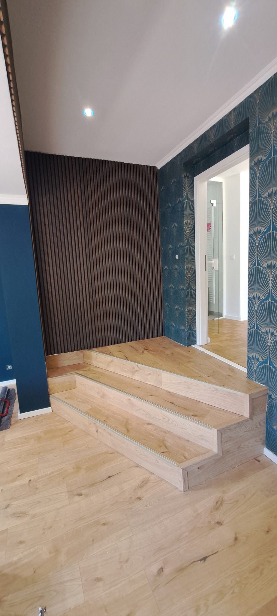 Nowoczesne wnętrze z drewnianymi schodami, panelami ściennymi i tapetą w stylu art deco. Widok na białe drzwi i dalsze pomieszczenie z drewnianą podłogą.