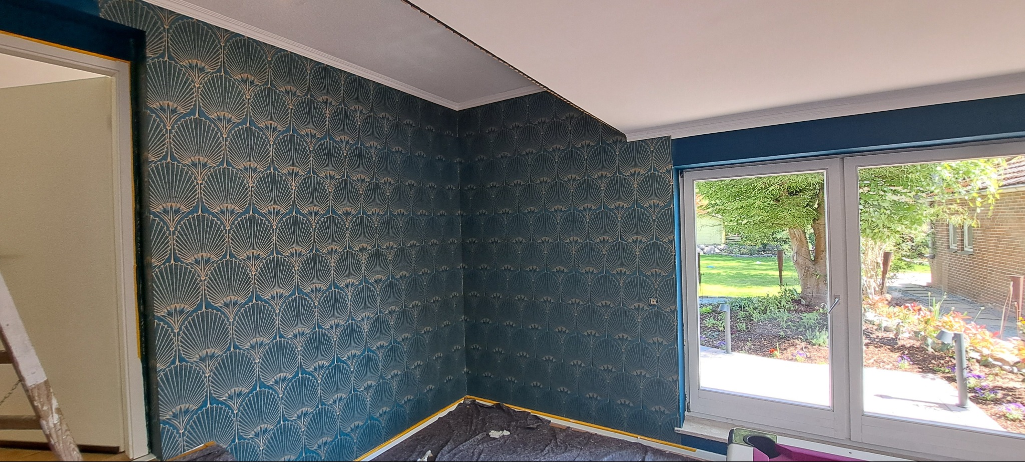 Ściana z tapetą w odcieniach niebieskiego i złota z motywem wachlarzy, widok na okno z ogrodem i częściowo odsłonięty sufit w narożniku pokoju.