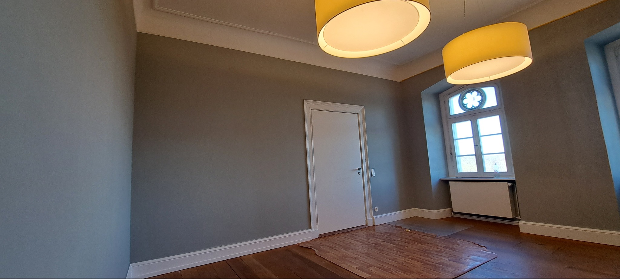 Pokój po remoncie: szare ściany, białe listwy przypodłogowe, drewniana podłoga, białe drzwi i okno z dekoracyjnym elementem. Dwie żółte lampy wiszące.