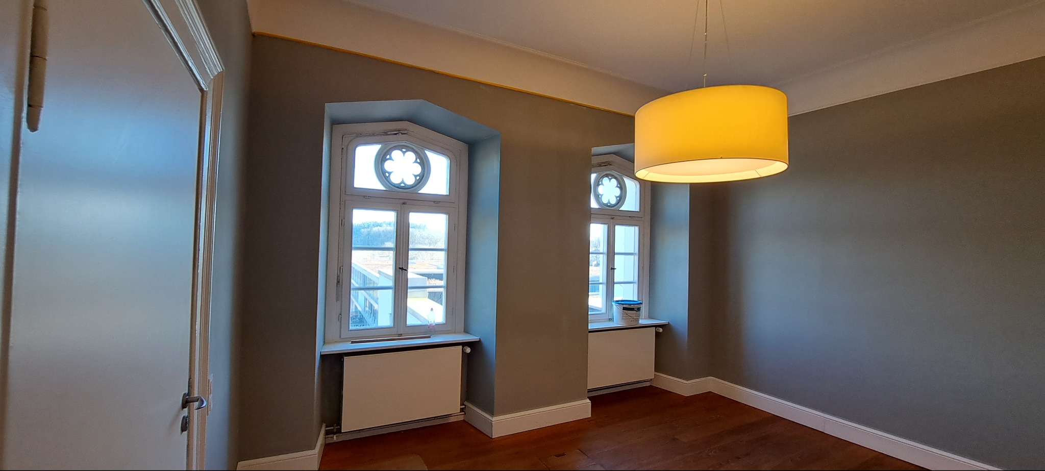 Wnętrze po malowaniu w Morągu: szare ściany, drewniana podłoga, okna z dekoracyjnym szprosowaniem i wisząca lampa. Na parapecie puszka farby.