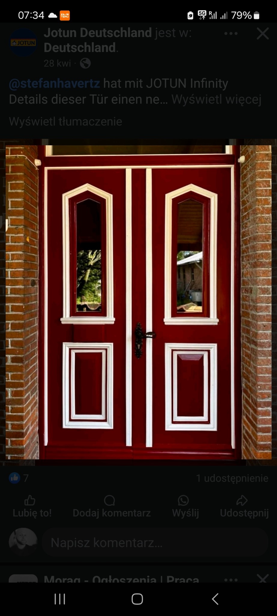 Czerwone drzwi wejściowe z białymi zdobieniami, widoczne przez szyby otoczenie domu, obok mur z czerwonej cegły, detale wykończenia.