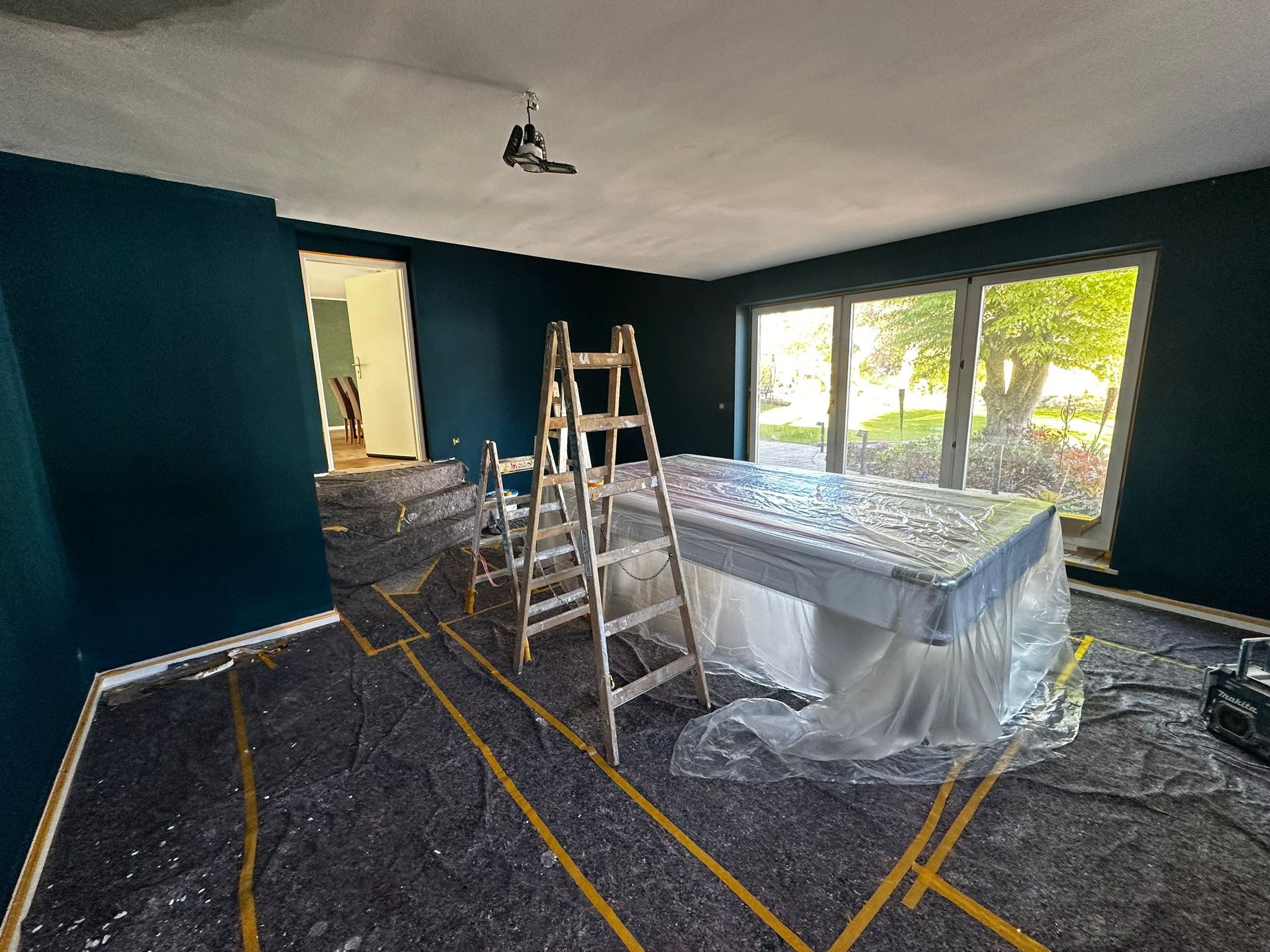 Pokój po malowaniu ścian na ciemny turkus, zabezpieczone meble i podłoga, widoczne drabiny malarskie. Widok na ogród przez okno.