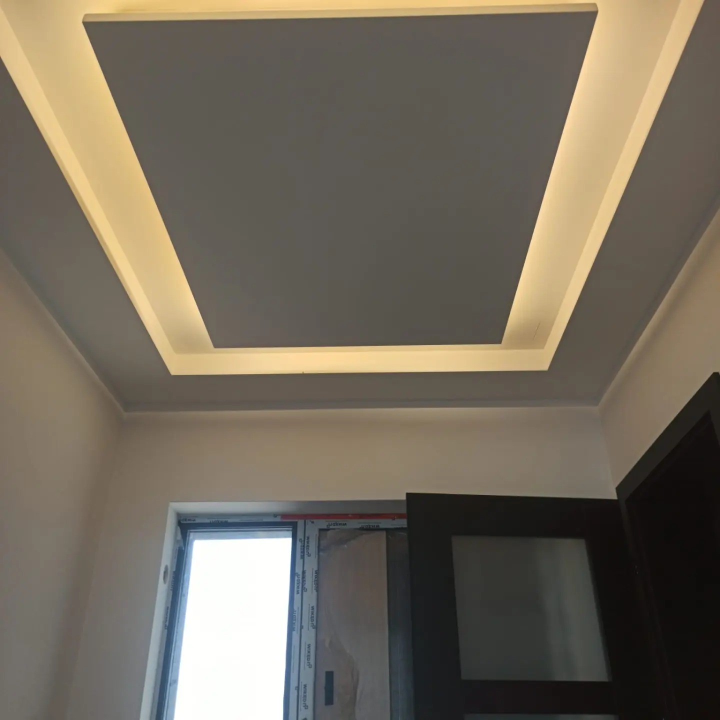 Szary sufit podwieszany z oświetleniem LED wzdłuż krawędzi, widoczny fragment okna i drzwi w wykończonym wnętrzu. Nowoczesny design sufitu.