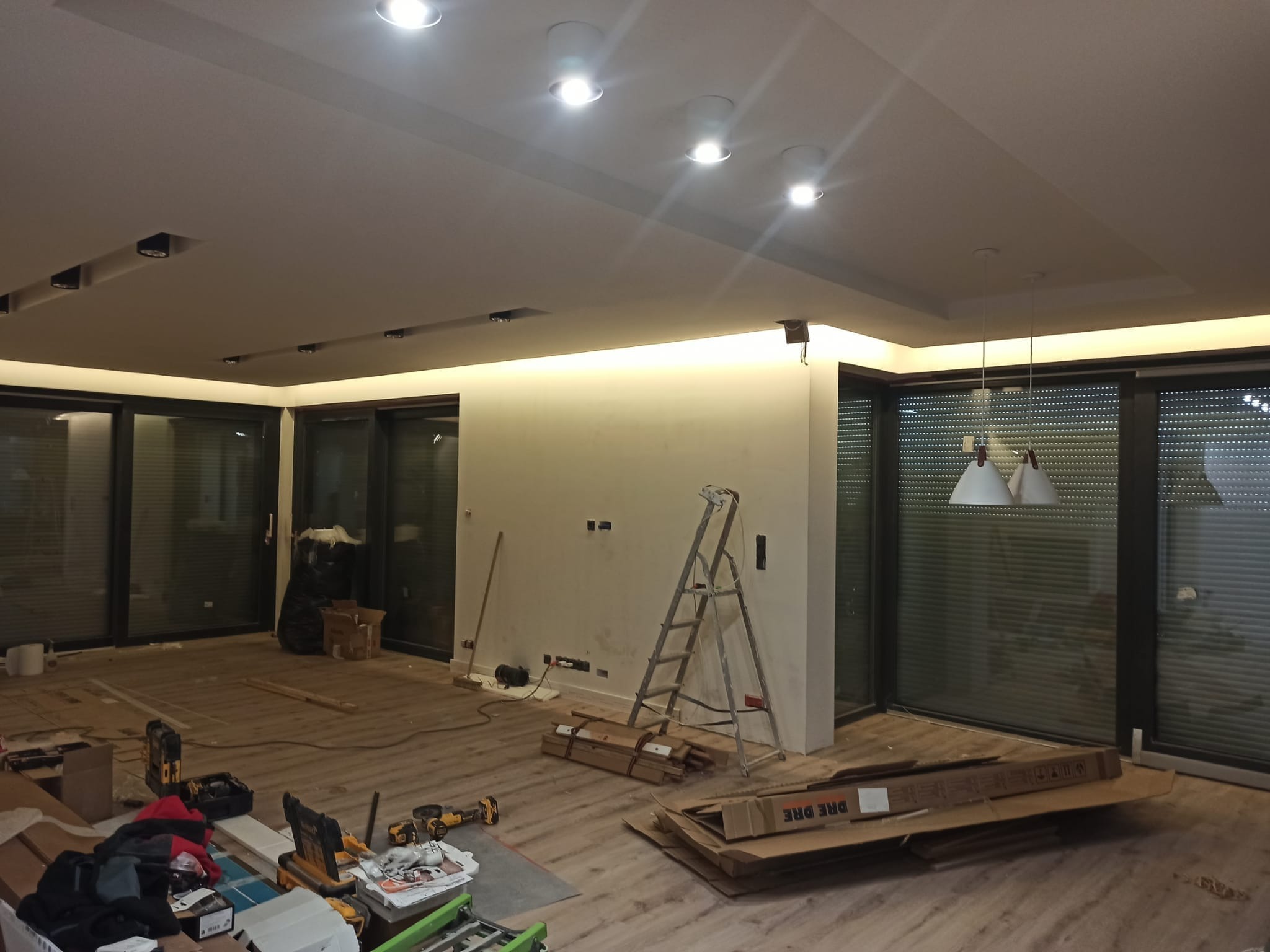 Wnętrze w trakcie prac wykończeniowych: widoczne narzędzia, drabina, podłoga częściowo zakryta, ściany przygotowane do malowania, oświetlenie sufitowe.