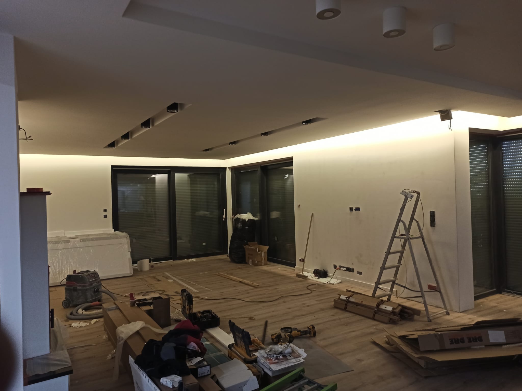 Wnętrze w trakcie prac wykończeniowych: surowe ściany, podłoga z desek, drabina, narzędzia i materiały budowlane. Widoczne oświetlenie sufitowe.