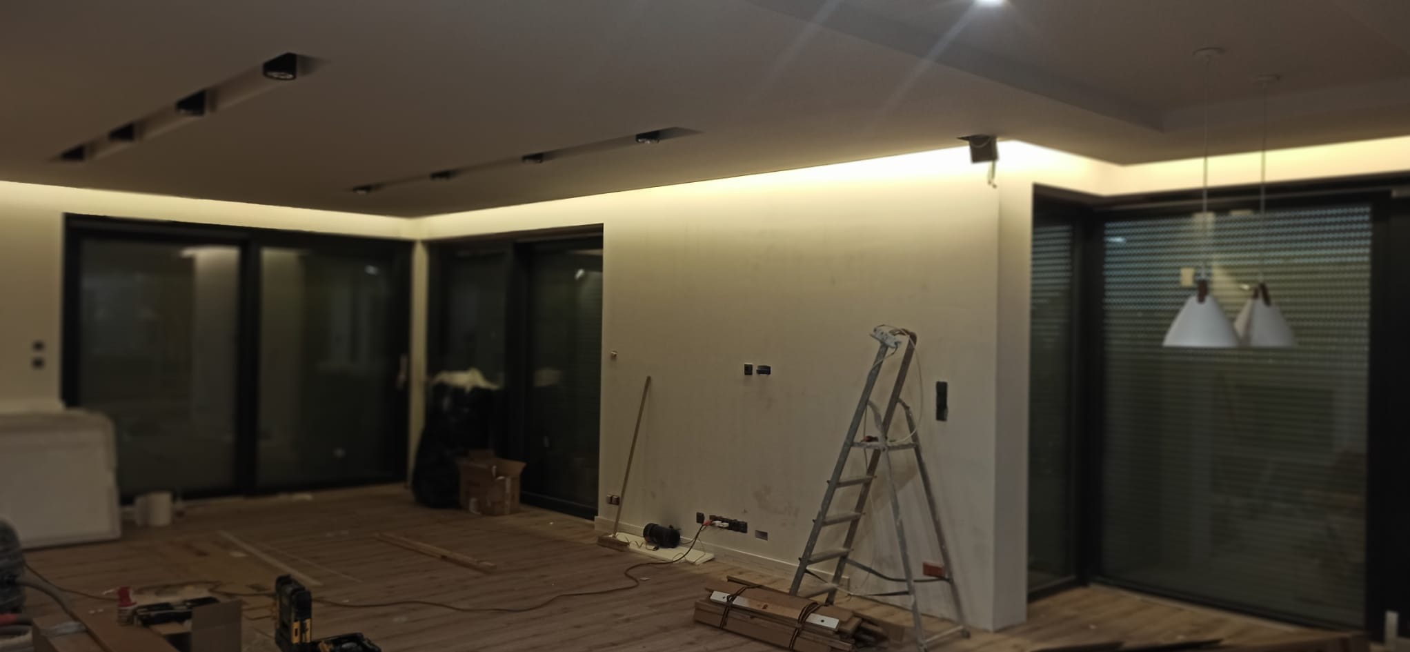 Wnętrze w trakcie wykończenia: surowa podłoga, ściany przygotowane do malowania, oświetlenie LED podsufitowe, drabina malarska. Widoczne gniazdka elektryczne.