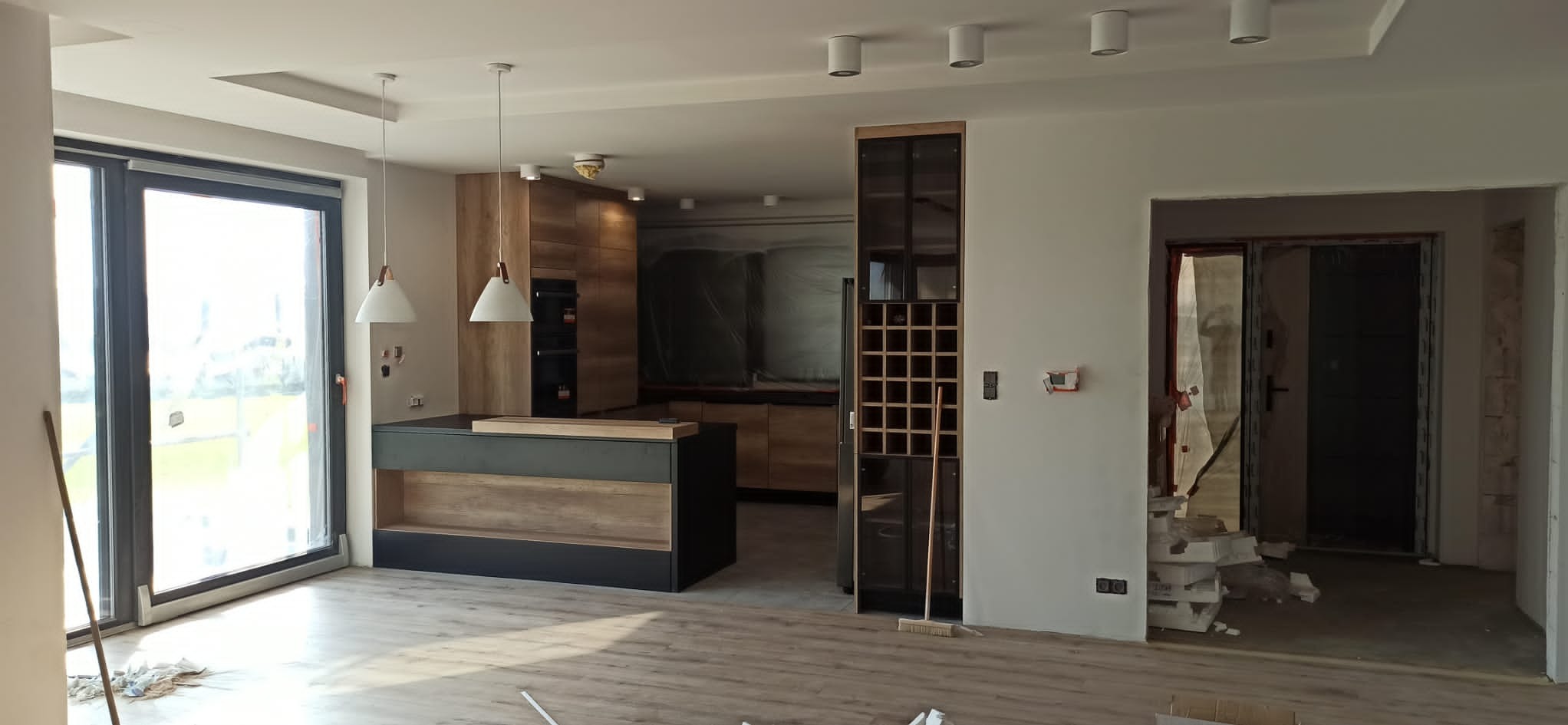 Jasne wnętrze domu w trakcie wykańczania: drewniana zabudowa kuchenna, wyspa, duże okno i drzwi balkonowe. Widoczne elementy instalacji elektrycznej.