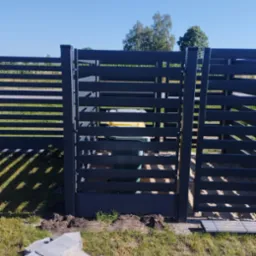 Nowoczesne, poziome ogrodzenie metalowe w kolorze grafitowym z furtką, z widocznym koszem na śmieci wewnątrz. Na pierwszym planie kostka brukowa.