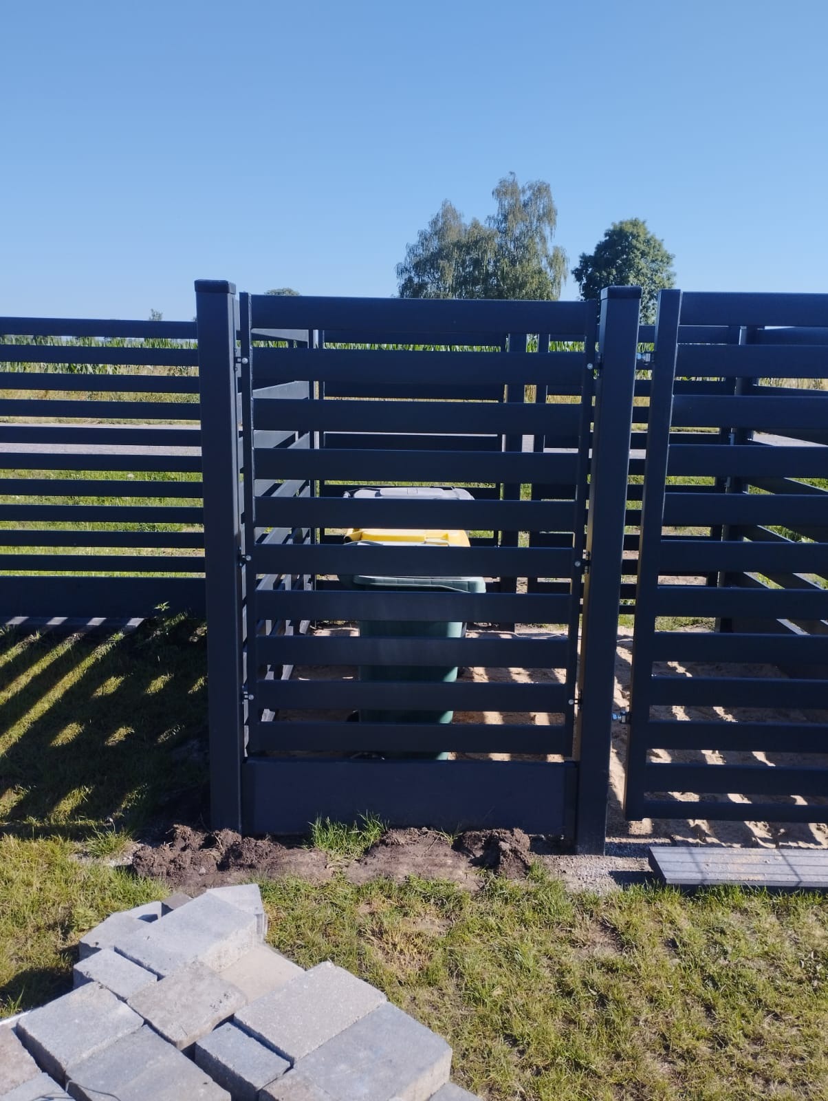 Nowoczesne, poziome ogrodzenie metalowe w kolorze grafitowym z furtką, z widocznym koszem na śmieci wewnątrz. Na pierwszym planie kostka brukowa.
