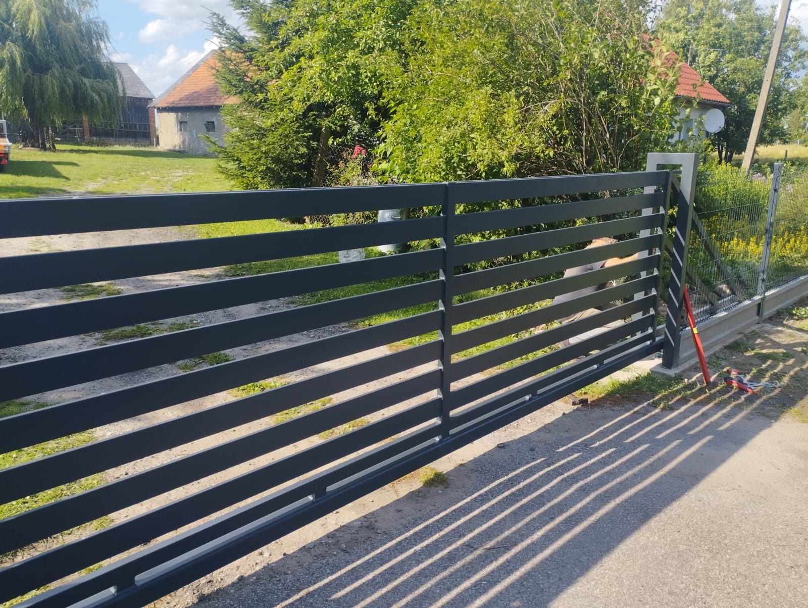 Nowoczesna, pozioma brama wjazdowa z metalowymi przęsłami w kolorze grafitowym, z widocznymi cieniami na podjeździe i zielenią w tle.