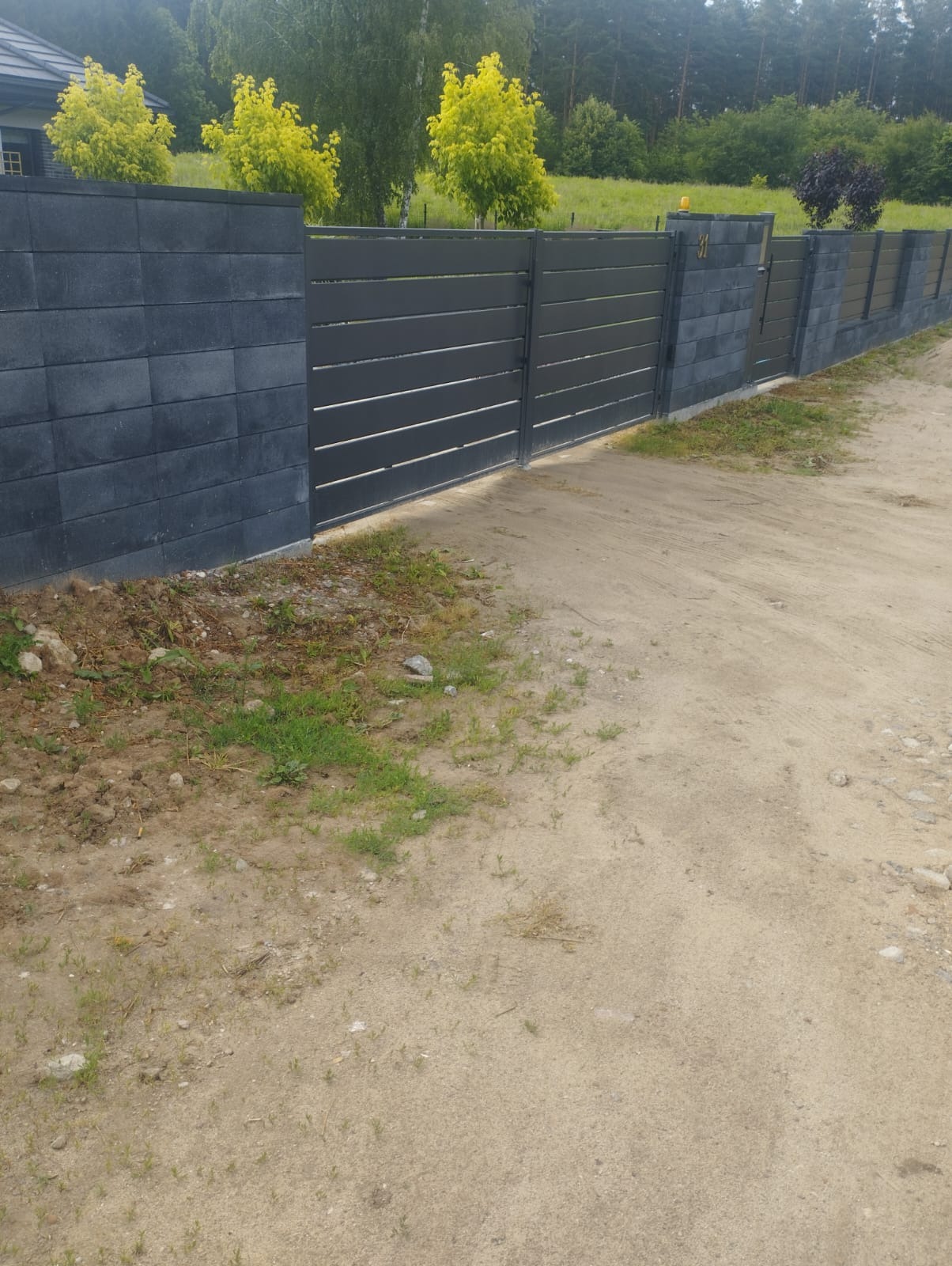 Nowoczesna, przesuwna brama wjazdowa z poziomymi panelami, osadzona w murze z szarej kostki. Widok na posesję z drzewami w tle, numer domu na murze.