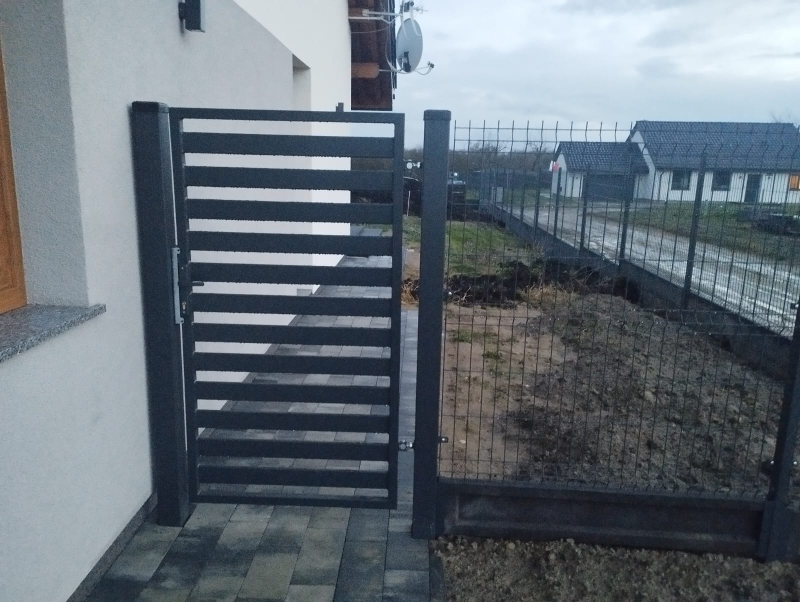 Nowoczesna, pozioma brama lamelowa w kolorze grafitowym, sąsiadująca z fragmentem ogrodzenia z siatki. Widok na posesję z domem w tle, pochmurne niebo.
