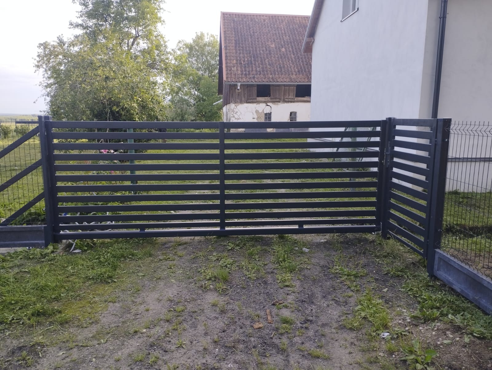 Nowoczesna, pozioma brama metalowa w Lubominie, z elementami siatki ogrodzeniowej. Widok na posesję z domem w tle, w pochmurny dzień.