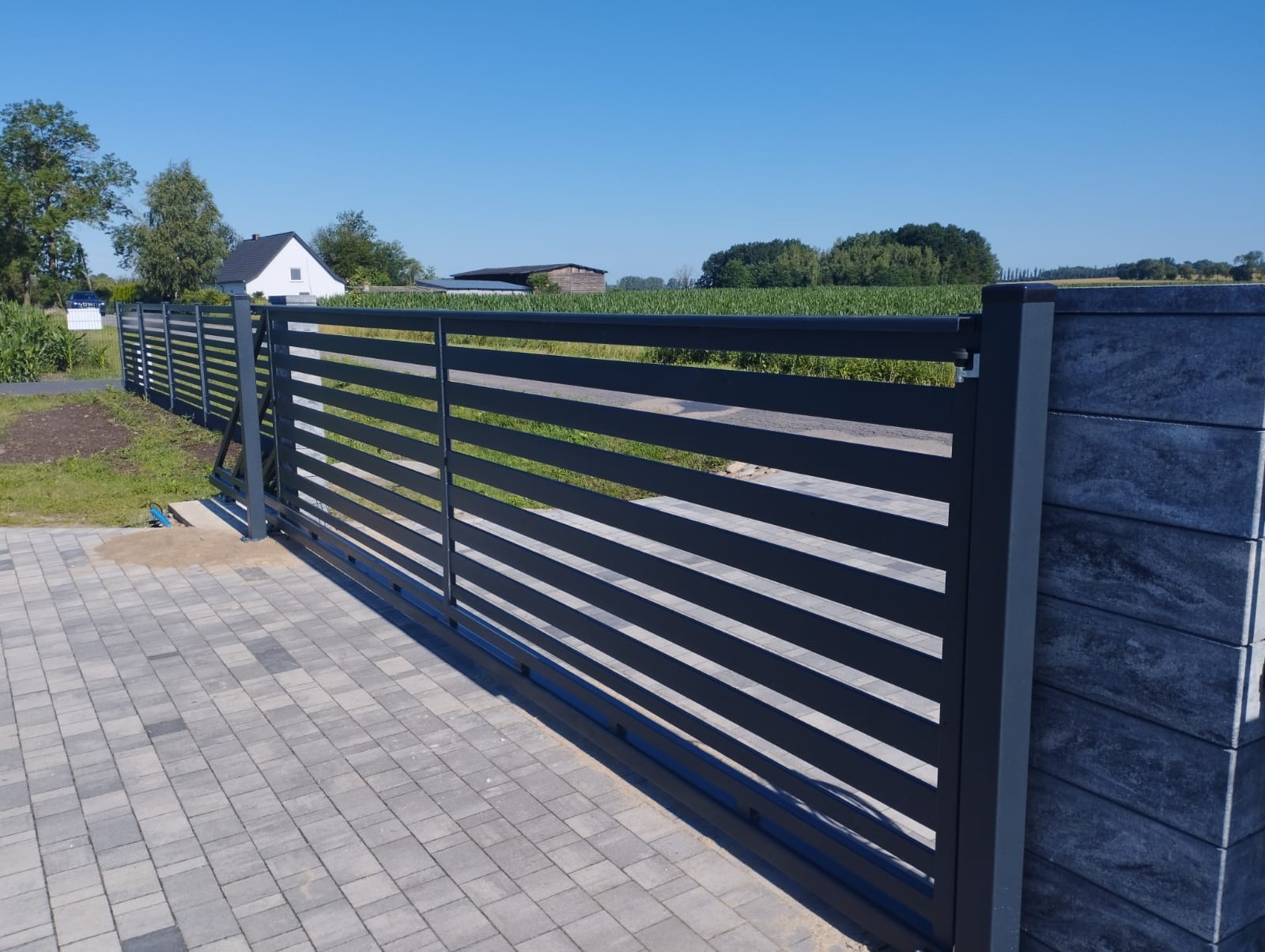 Nowoczesna, przesuwna brama wjazdowa z poziomymi panelami, szara kolorystyka, kostka brukowa na podjeździe, widok na zielone pole i dom w tle.