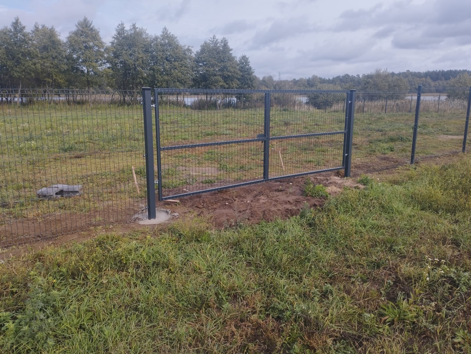 Szara, dwuskrzydłowa brama panelowa zamontowana w ogrodzeniu z siatki na tle krajobrazu z drzewami i zbiornikiem wodnym w oddali. Widoczne świeże fundamenty słupków.