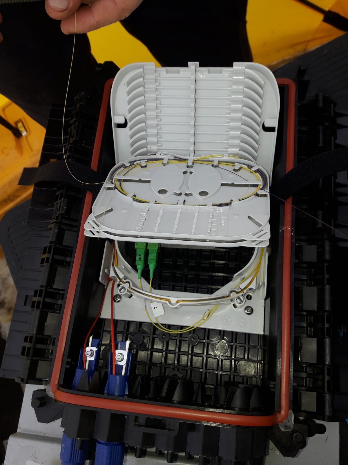 Otwarta skrzynka telekomunikacyjna z widocznymi kablami światłowodowymi i złączami, w trakcie instalacji lub naprawy. Widoczne narzędzia i elementy montażowe.