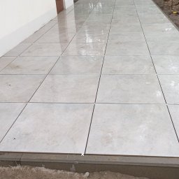 Solid-Bruk Błażej Sikorski - Ułożona jasna, kwadratowa kostka brukowa przy ścianie budynku z widocznymi krzyżykami dystansowymi i obrzeżem, na przygotowanym podłożu.