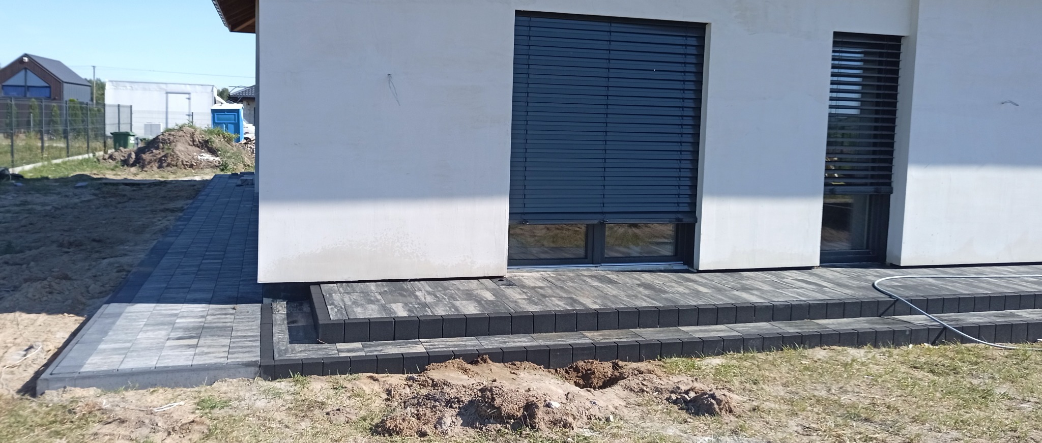 Ułożona kostka brukowa w odcieniach szarości tworzy schody i ścieżkę przy białym budynku z oknami. Widoczny fragment ogrodu i domu w tle.