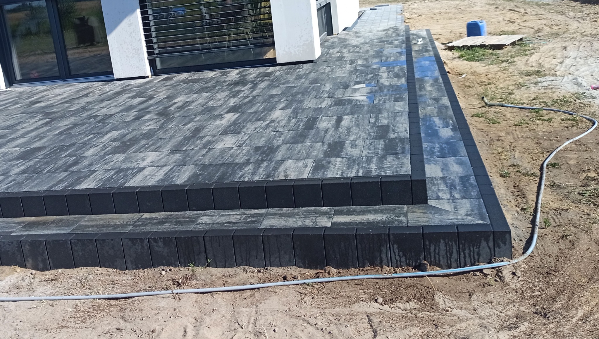 Nowoczesny taras z kostki brukowej o szaro-białej kolorystyce, wykończony czarnym obrzeżem. Widoczny fragment budynku z oknem i niezabudowany teren wokół.