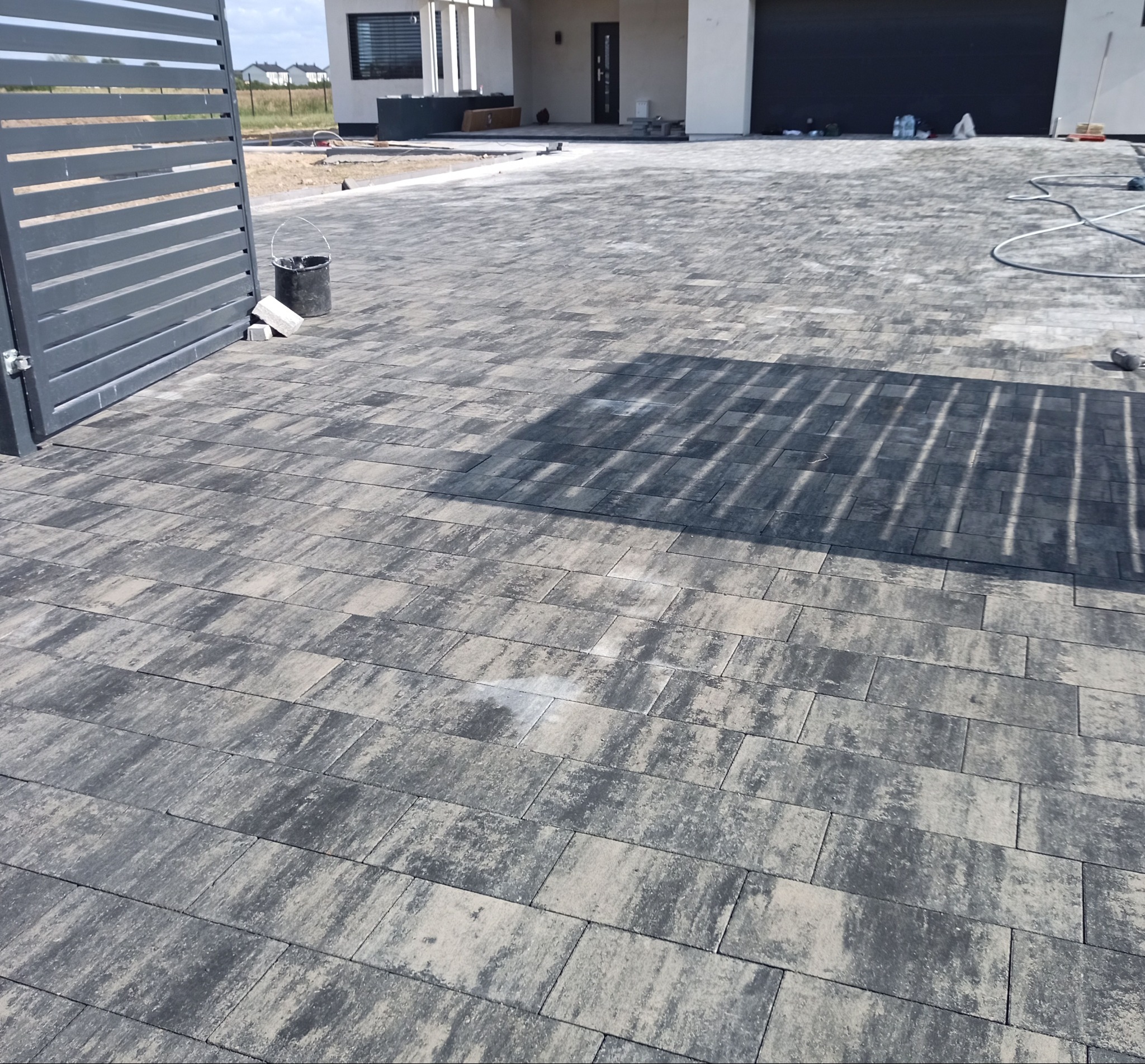 Ułożona kostka brukowa w odcieniach szarości prowadząca do nowoczesnego domu z garażem. Widoczny fragment ogrodzenia i wiadro.