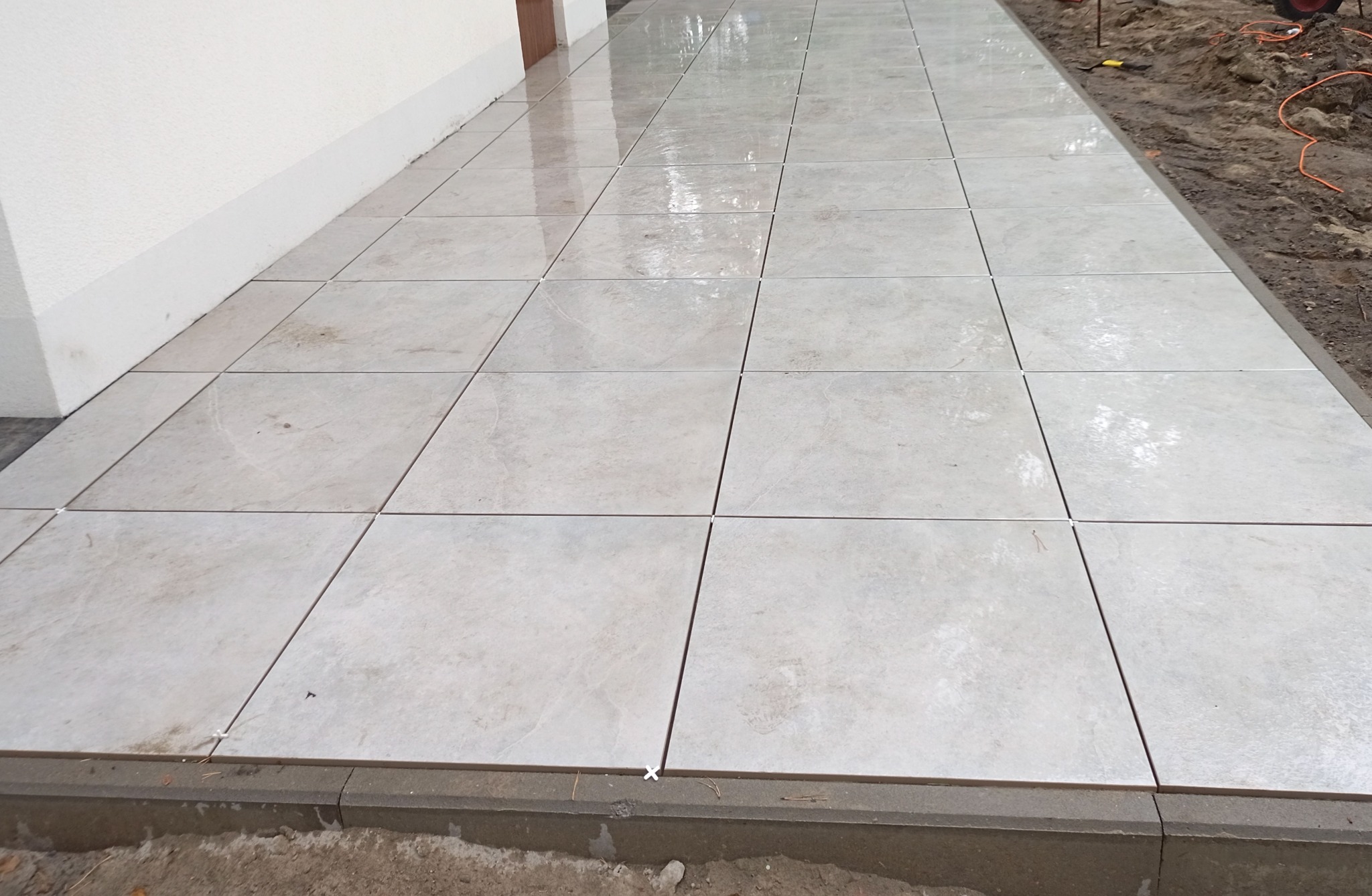 Ułożona jasna, kwadratowa kostka brukowa przy ścianie budynku z widocznymi krzyżykami dystansowymi i obrzeżem, na przygotowanym podłożu.