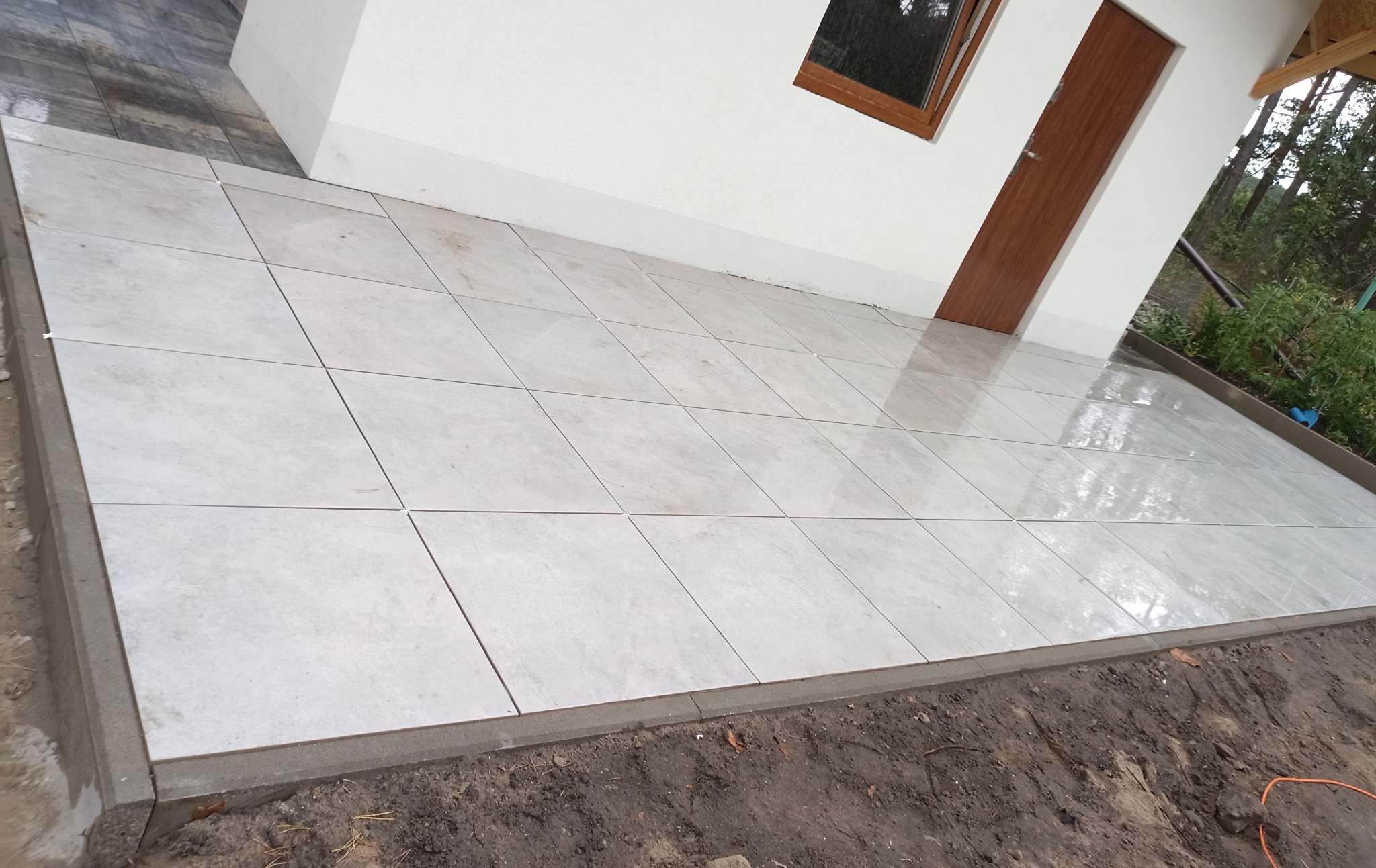 Jasne, kwadratowe płytki ułożone na zewnątrz budynku z brązowymi drzwiami i oknem, otoczone zielenią. Widoczne krawędzie z betonu.