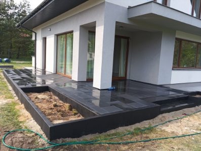 Nowoczesny taras z ciemnej kostki granitowej przy białym domu. Widoczna rabata z ziemią, schody tarasowe i odbicie w mokrej powierzchni kostki.