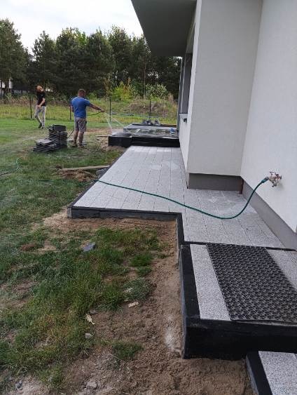 Jasnoszara kostka brukowa wokół domu, myta wodą z węża przez mężczyznę w niebieskiej koszulce. Fragment trawnika i czarne obrzeża kostki.