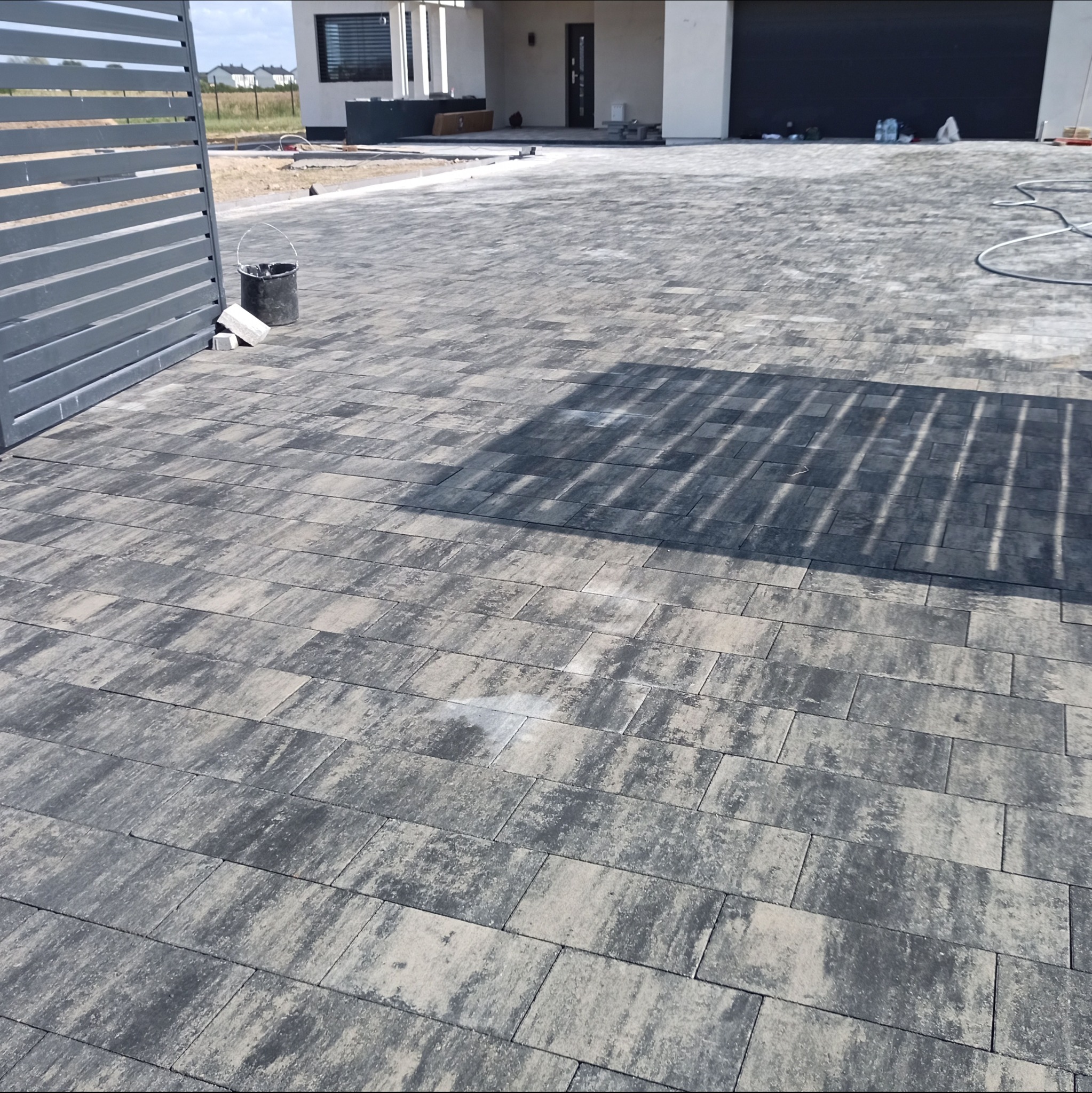 Ułożona szara kostka brukowa na podjeździe przed domem z widocznym fragmentem szarego ogrodzenia i elementami budowy w tle.