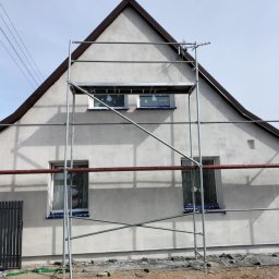 Robert Dementow - Elewacja domu jednorodzinnego w trakcie prac wykończeniowych. Widoczne rusztowanie, okna zabezpieczone folią, świeżo nałożony tynk. Ujęcie z dołu.