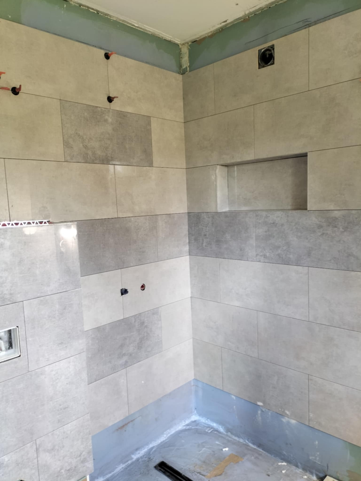 Łazienka w trakcie remontu: szare, prostokątne płytki na ścianach, widoczny odpływ liniowy w podłodze, wnęka w ścianie, elementy instalacji.