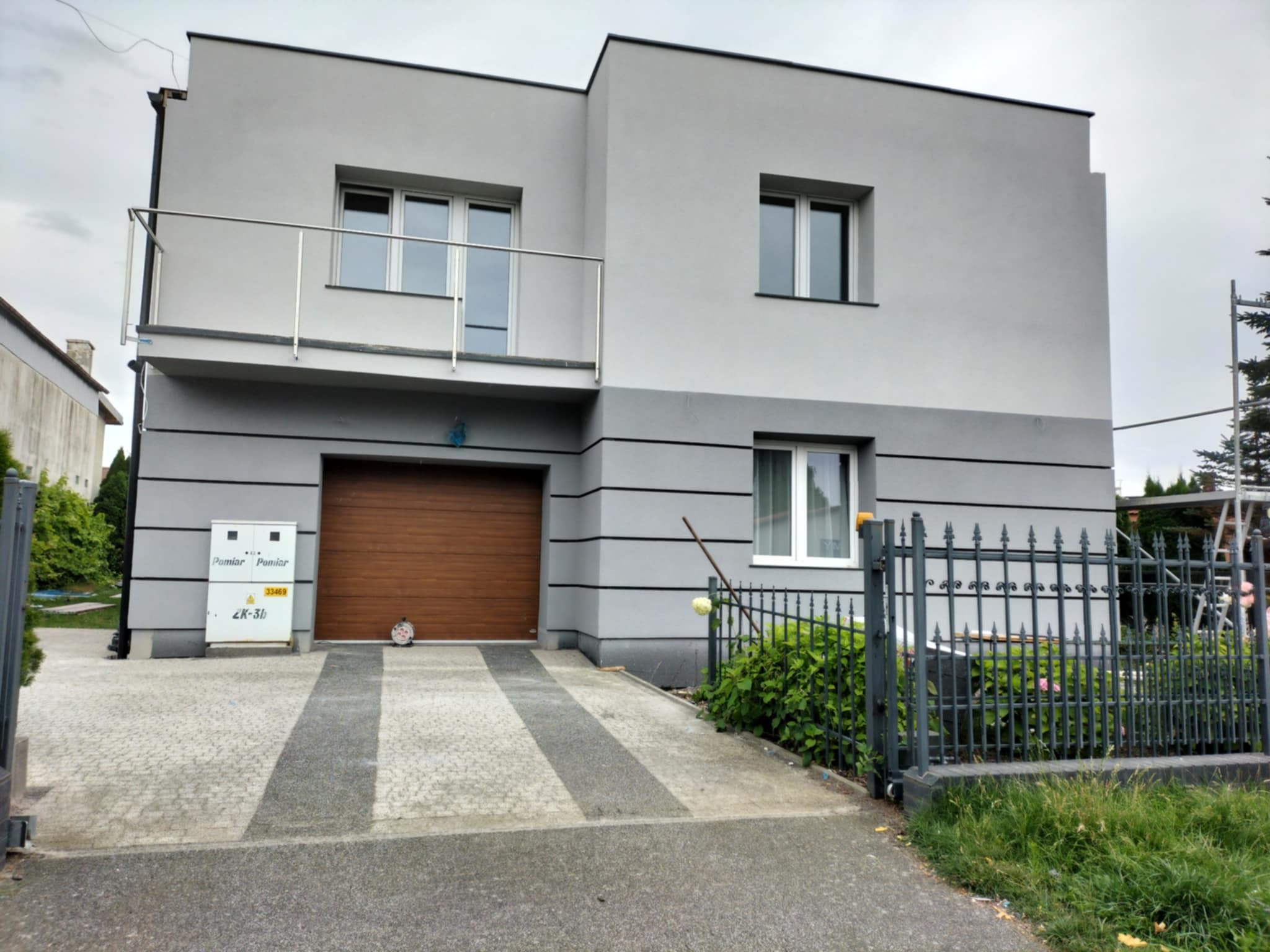 Dwukondygnacyjny dom z szarą elewacją, brązową bramą garażową i balkonem z metalową balustradą. Widoczny fragment ogrodzenia i chodnika z kostki brukowej.