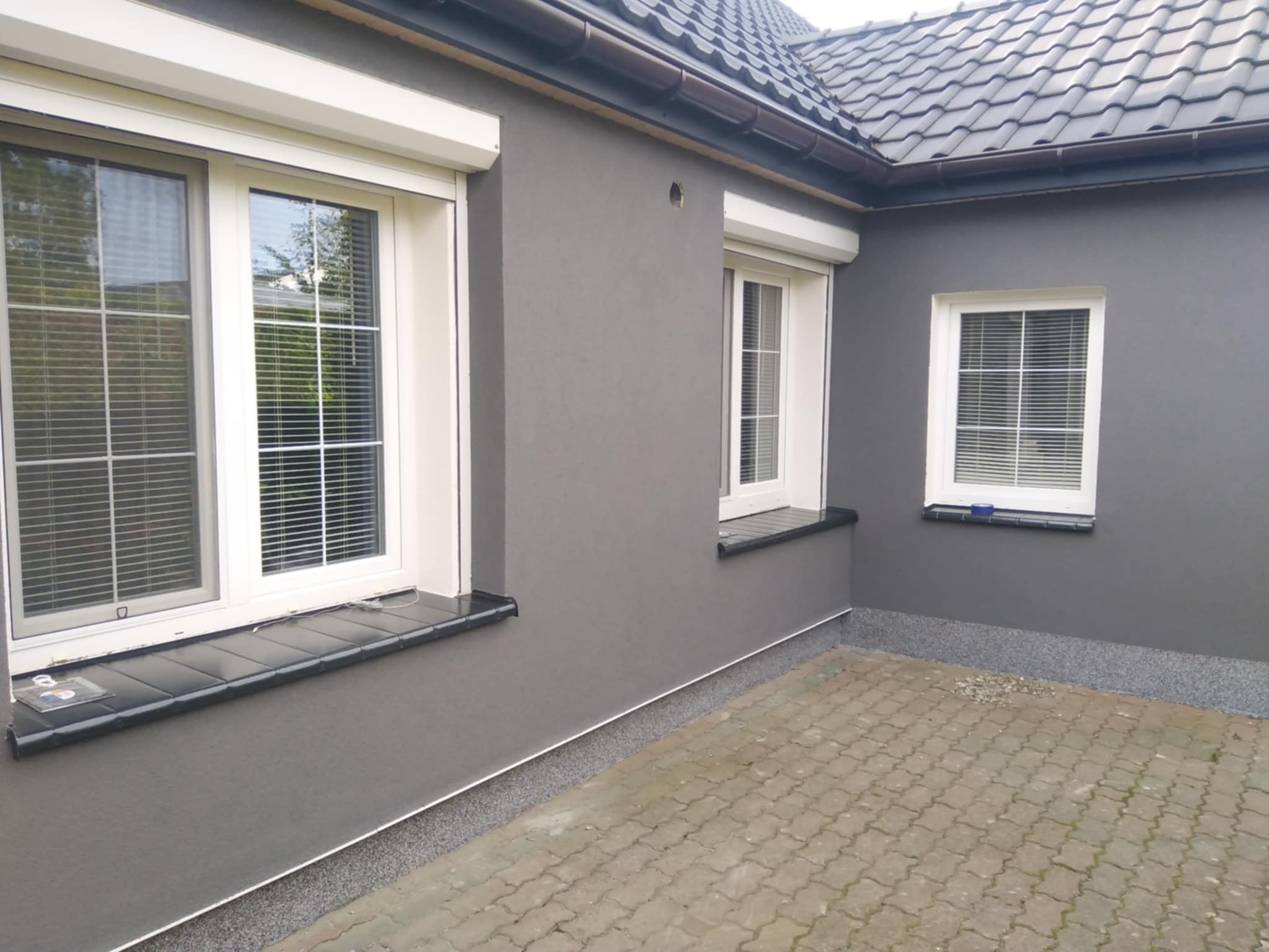 Szara elewacja domu jednorodzinnego z trzema oknami z roletami zewnętrznymi, wykończona ciemną podmurówką i parapetami z płytek. Widoczna kostka brukowa.