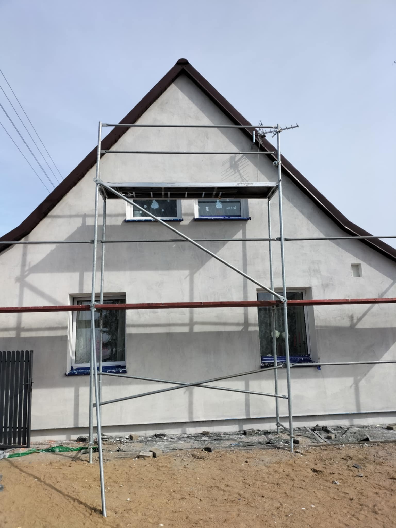 Elewacja domu jednorodzinnego w trakcie prac wykończeniowych. Widoczne rusztowanie, okna zabezpieczone folią, świeżo nałożony tynk. Ujęcie z dołu.